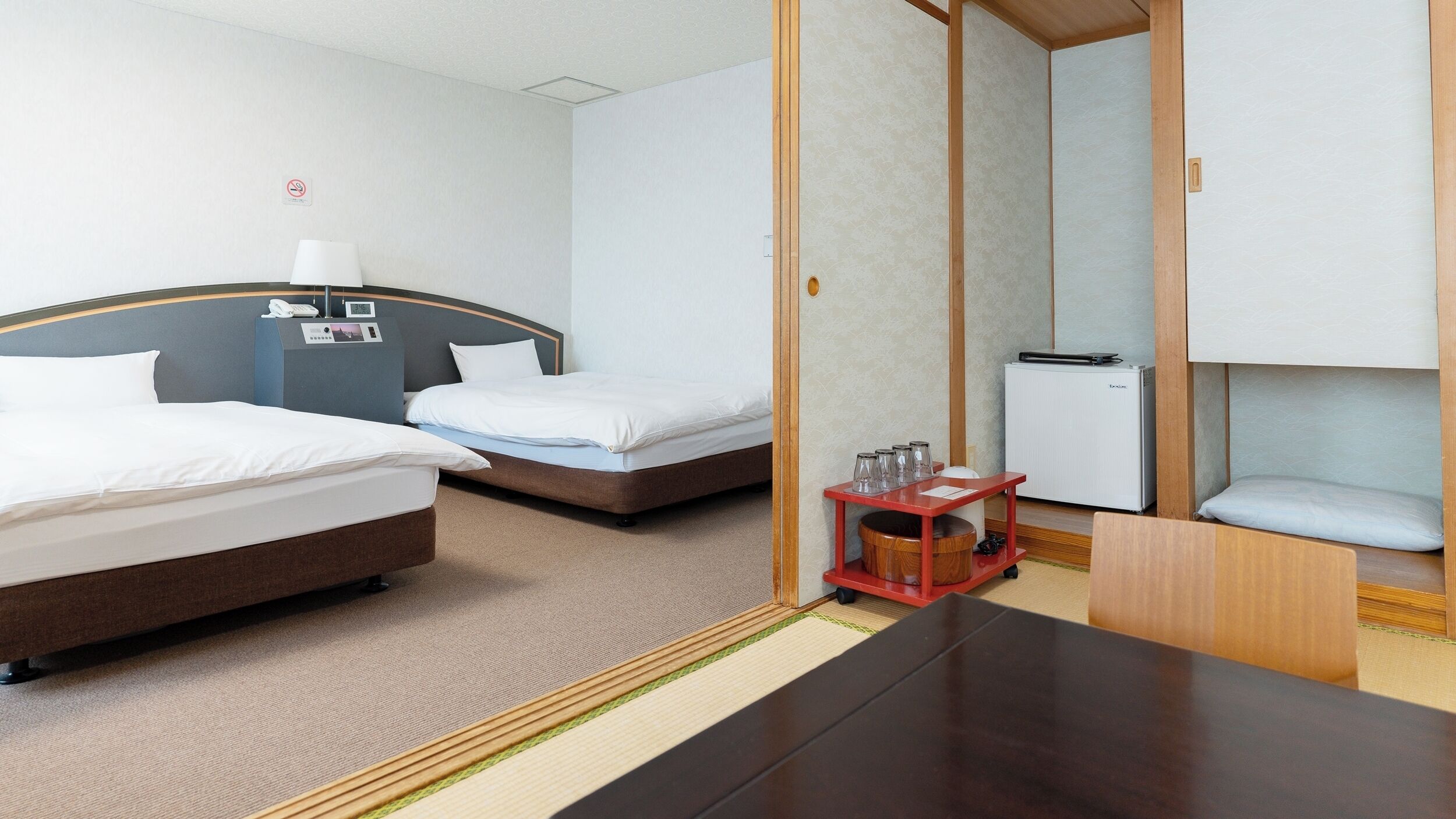 A hotel room with two beds, a desk, a minibar, and a small table with glasses.