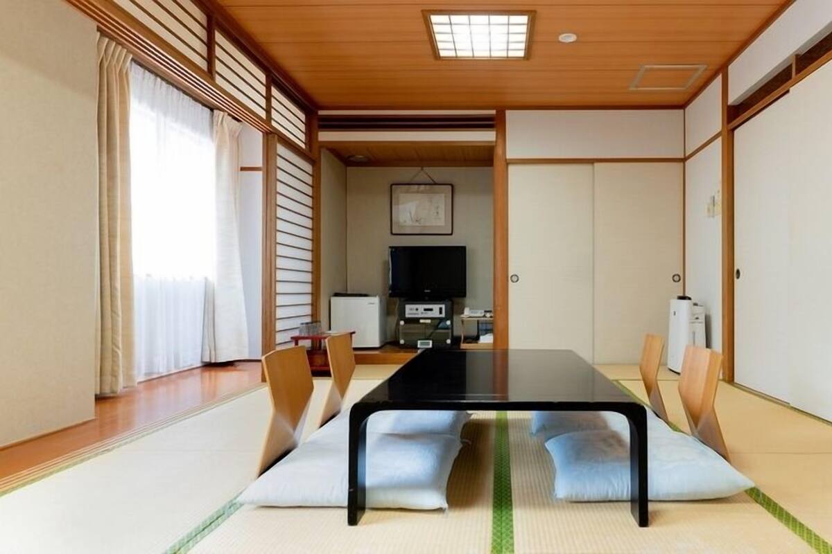 Une pièce avec un sol recouvert de tatamis, une petite table basse, des coussins, des portes coulissantes et un téléviseur.