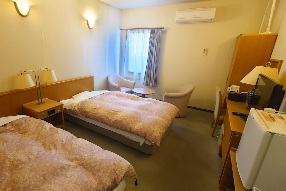 A hotel room with two beds, a desk, a chair, a small table, and an air conditioning unit.