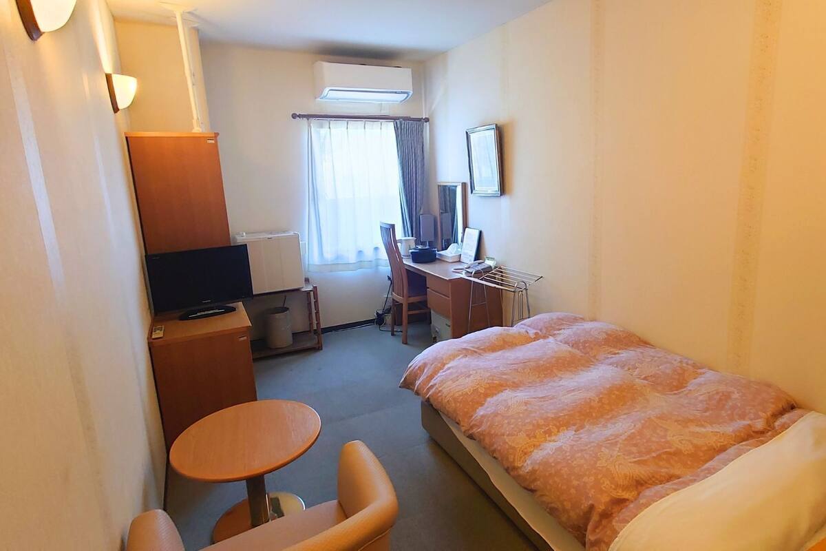 A hotel room with a bed, a desk with a computer, a chair, a TV, and a window with curtains.