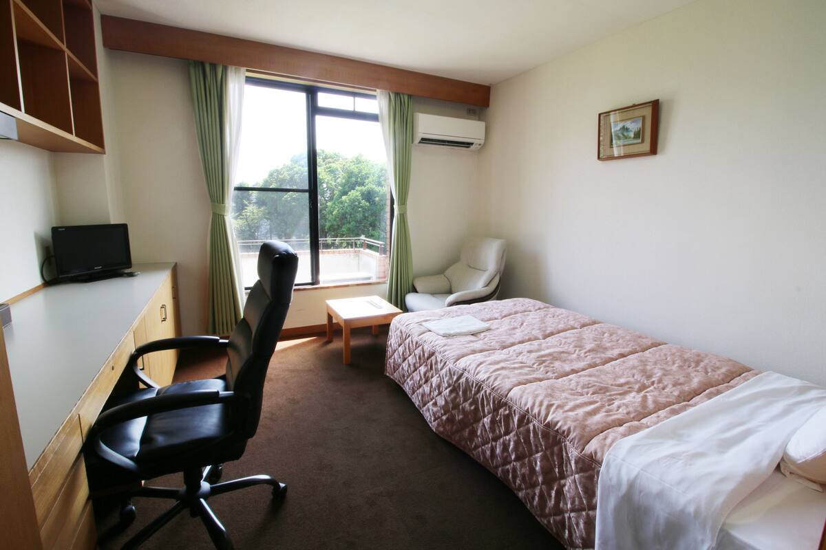 A hotel room with a bed, a desk with a computer, a chair, a window with curtains, and an air conditioning unit.
