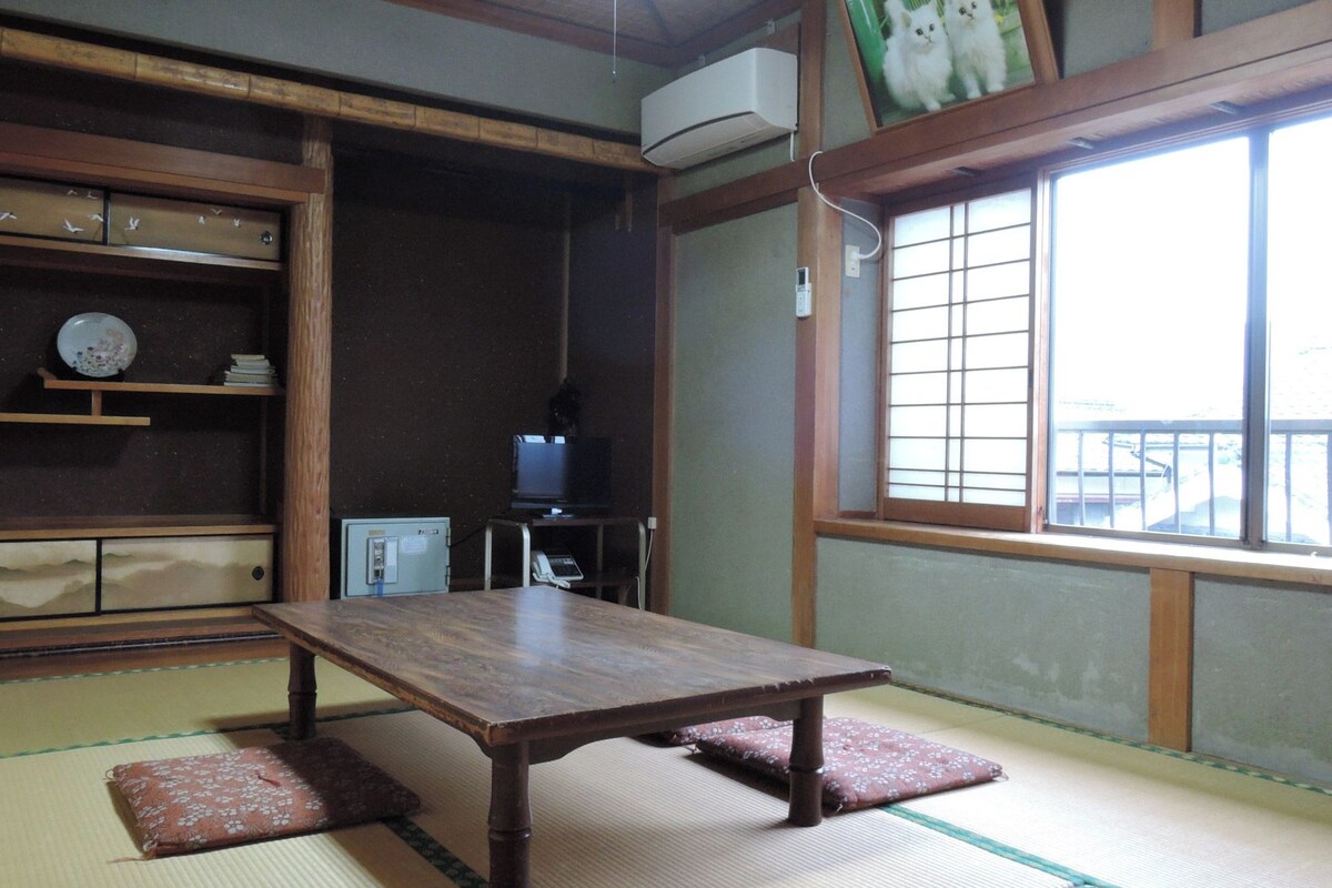 A traditional Japanese room with tatami flooring, a wooden table, a television, and a window with a view.
