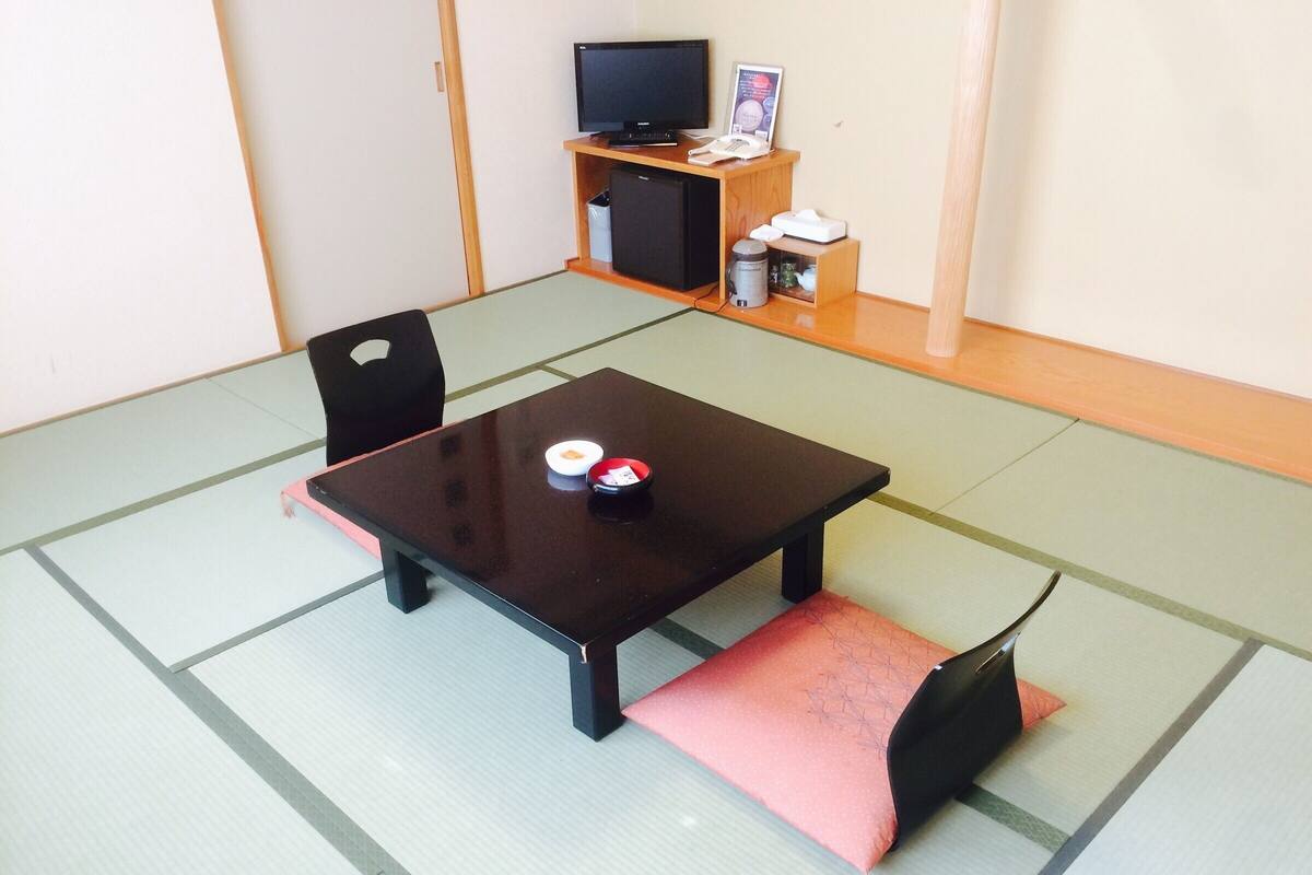 A tatami mat room with a low table, two chairs, a television, and a small shelf with items.