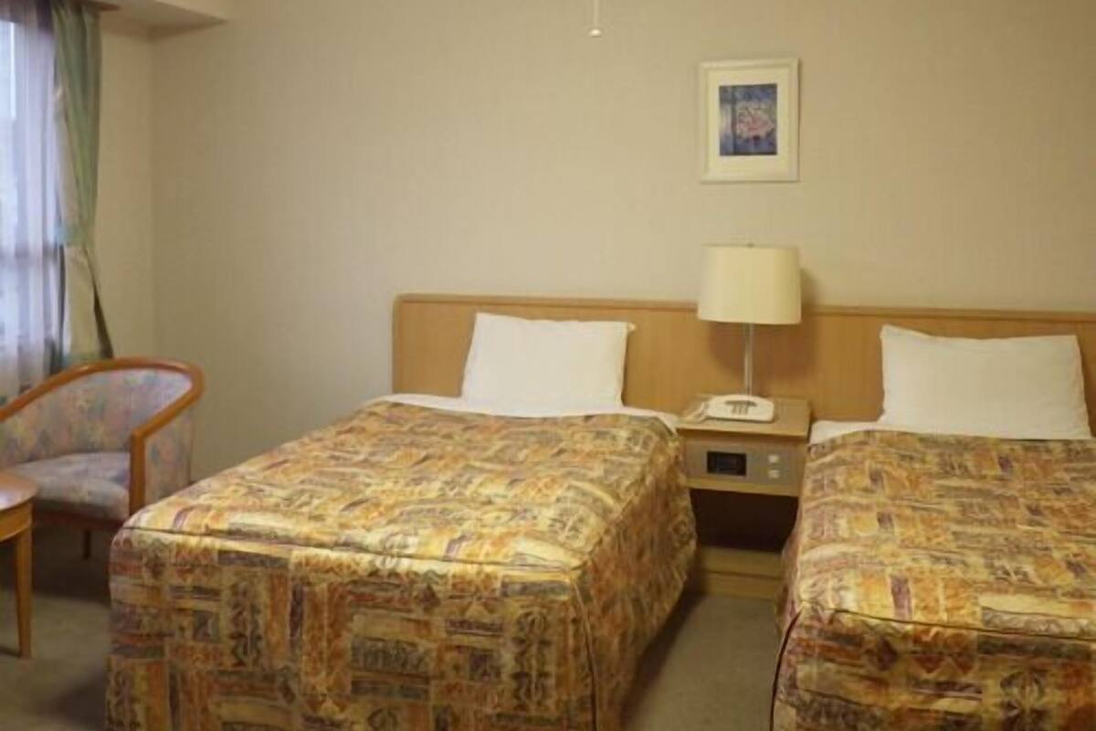 A hotel room with two beds, a chair, a nightstand with a lamp, and a framed picture on the wall.
