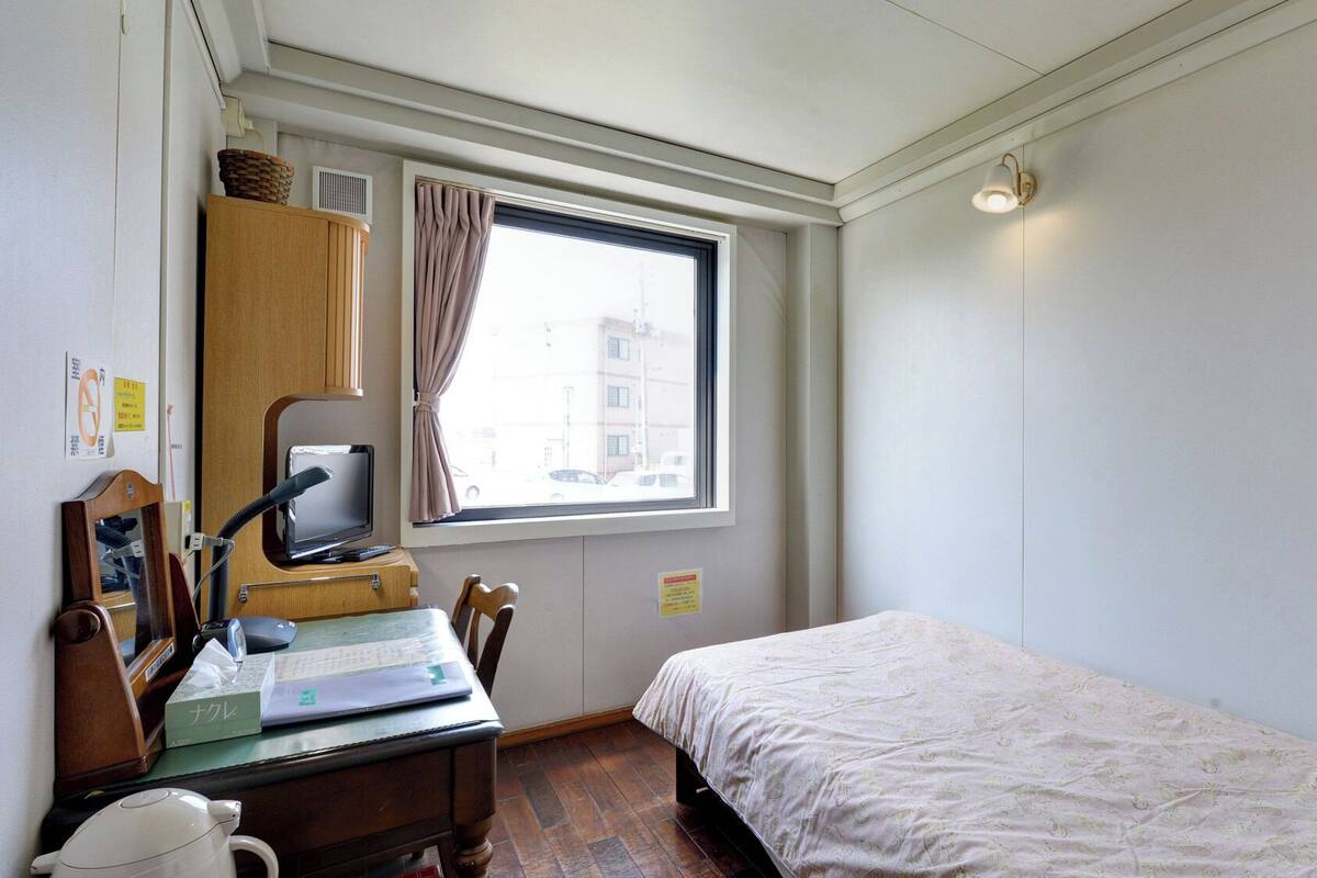 A small bedroom with a single bed, a desk with a computer, a mirror, and a window with curtains.