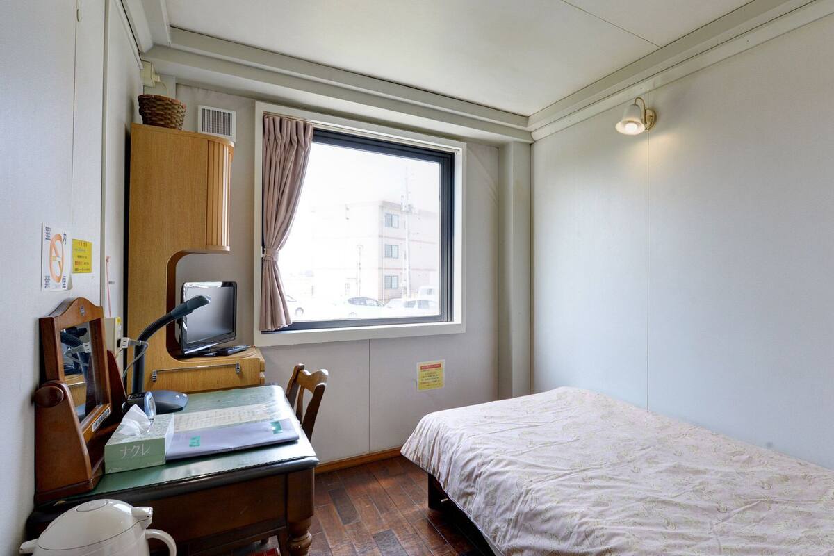 A small, tidy room with a bed, a desk with a computer, a mirror, and a window with curtains.