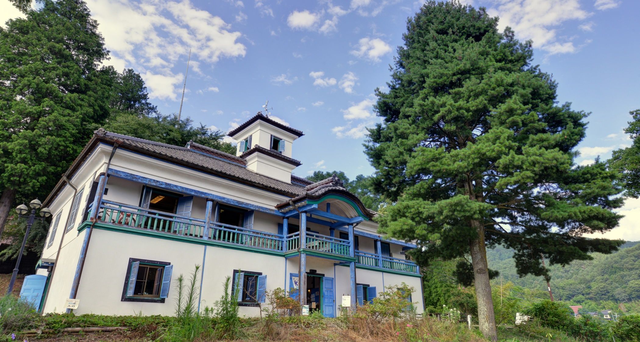 A traditional building with a balcony, surrounded by trees and a mountainous landscape.