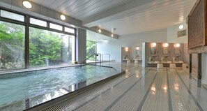 A modern indoor swimming pool with clear glass walls, a tiled floor, and a well-lit bathroom area with sinks and mirrors. - Oishii Gakko (Hokuto)