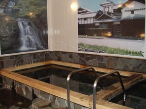 A traditional Japanese onsen with a wooden bath, stone walls, and a view of a waterfall and traditional buildings. - Hotel Tamai (Aki)