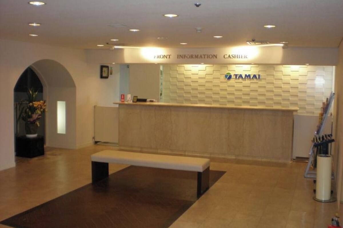 Un área de recepción con un banco, un mostrador con la etiqueta 'FRONT INFORMATION CASHIER' y una pared con el logotipo 'TAMAI'.