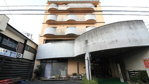 A multi-story building with a curved facade, a gated entrance, and a small green area in front. - Okumusashi Ryokan (Hanno)