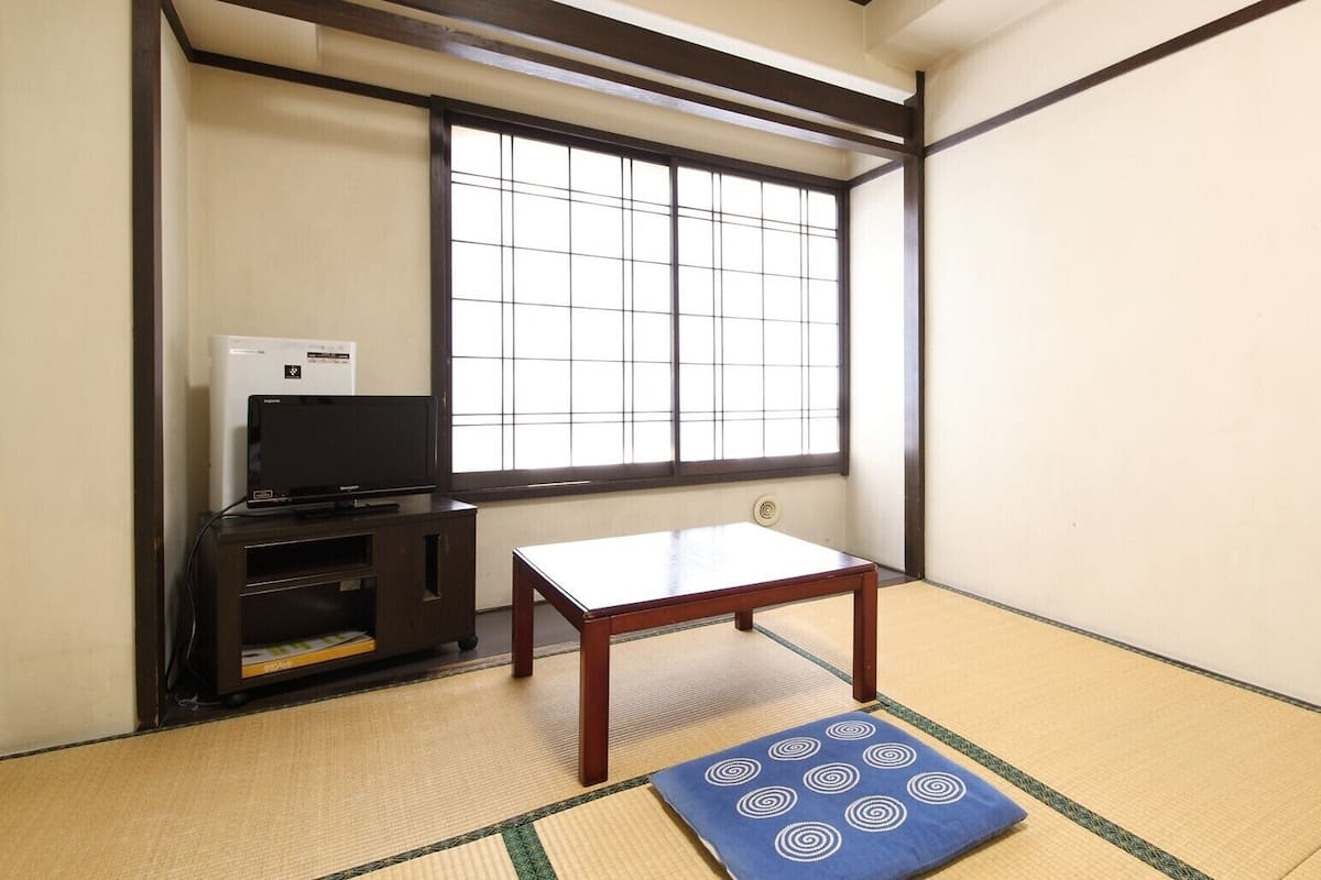 A traditional Japanese room with tatami flooring, a low wooden table, a television on a cabinet, and a large window with grid patterns.
