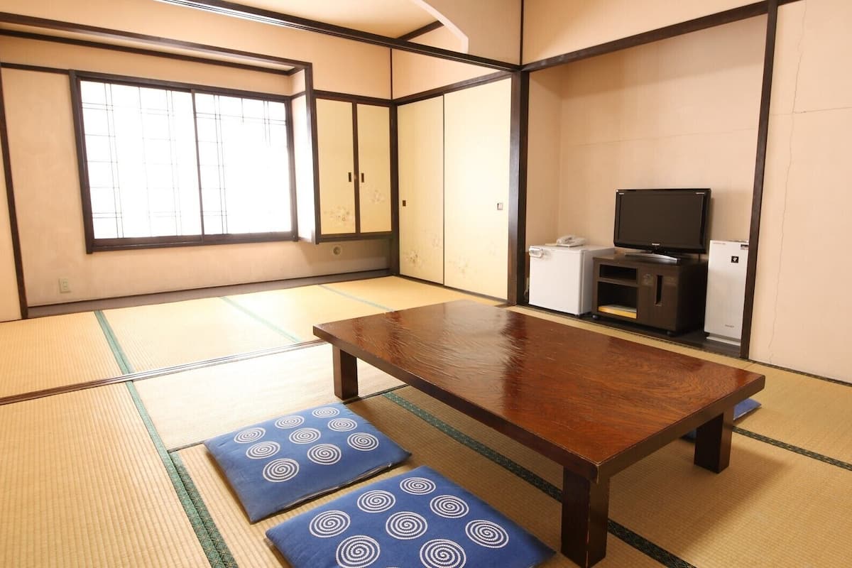A tatami mat room with a wooden table, cushions, a television, and a small refrigerator.