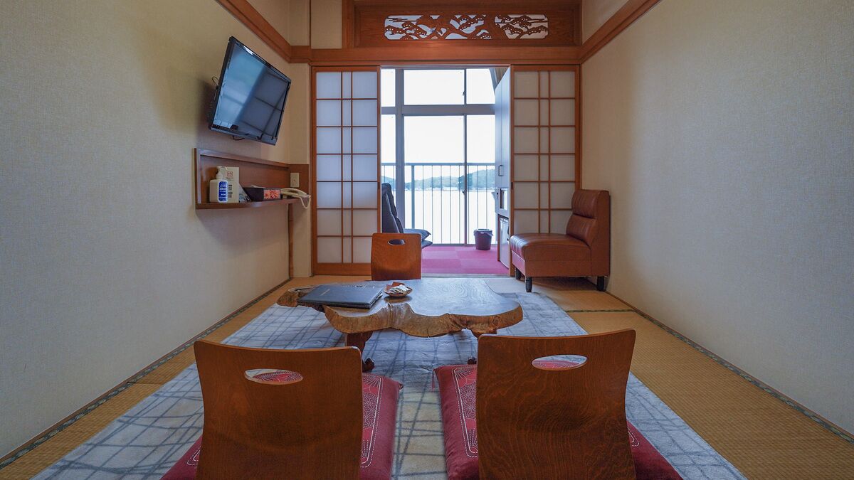 Ein traditioneller japanischer Raum mit Tatami-Böden, einem niedrigen Tisch, zwei hölzernen Stühlen, einem an der Wand montierten Fernseher und einer Schiebetür, die auf einen Balkon führt.