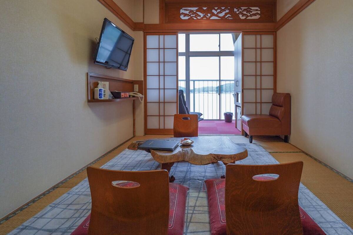 Ein traditioneller japanischer Raum mit Tatami-Böden, einem niedrigen Tisch, zwei hölzernen Stühlen, einem an der Wand montierten Fernseher und einer Schiebetür, die auf einen Balkon führt.