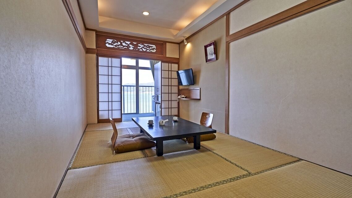 Ein traditioneller japanischer Raum mit Tatami-Boden, einem niedrigen Tisch, Kissen, einer Schiebetür und einem an der Wand befestigten Fernseher.