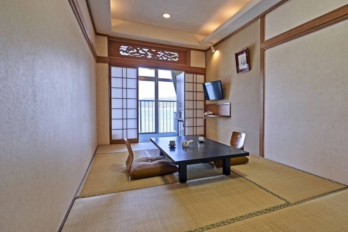 Ein traditioneller japanischer Raum mit Tatami-Boden, einem niedrigen Tisch, Kissen, einer Schiebetür und einem an der Wand befestigten Fernseher.