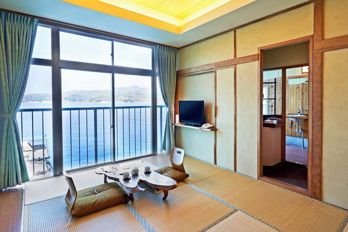 Ein Zimmer mit großem Fenster, Fernseher, kleinem Tisch mit Tassen und Meerblick.