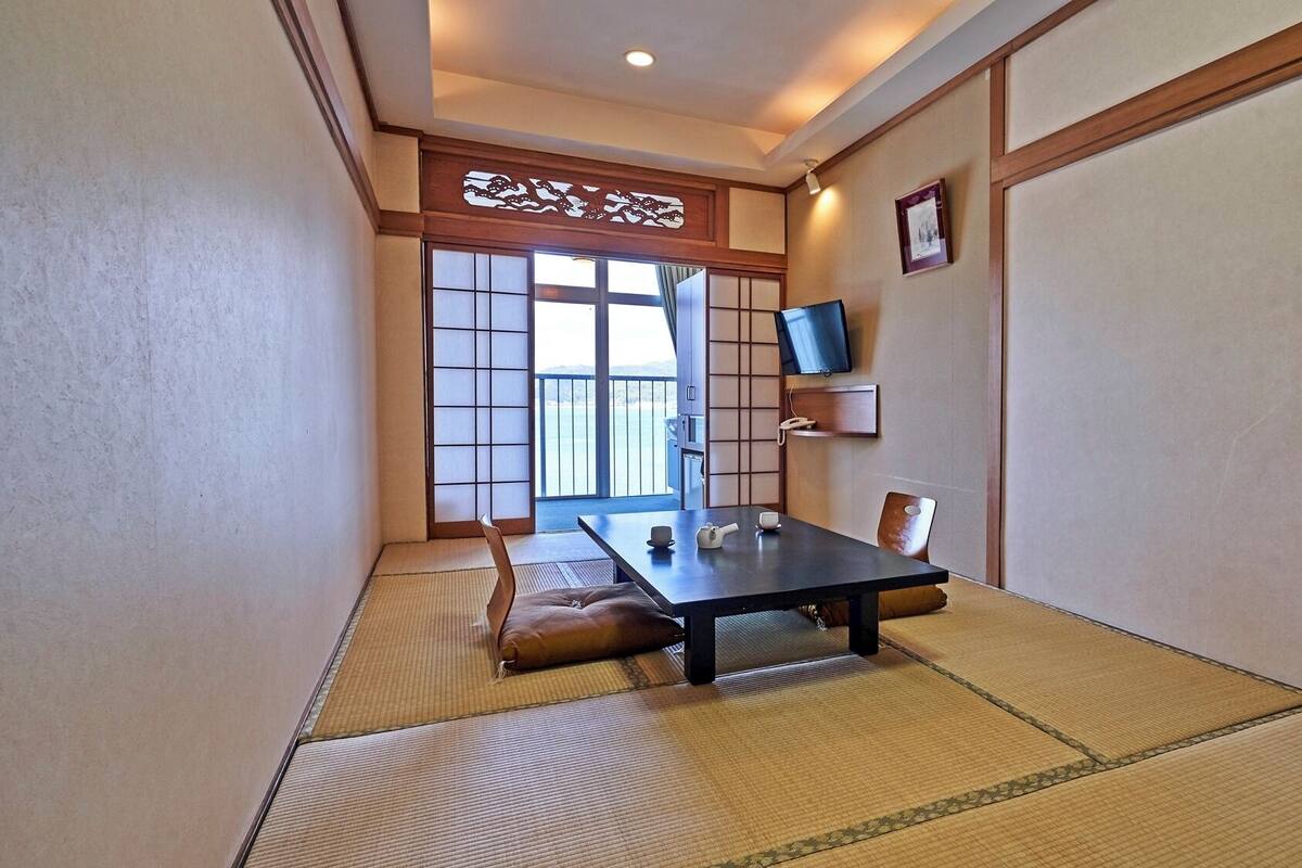 Ein traditioneller japanischer Raum mit Tatami-Bodenbelag, einem niedrigen Tisch mit Teeservice, einer Schiebetür mit Blick und einem an der Wand befestigten Fernseher.