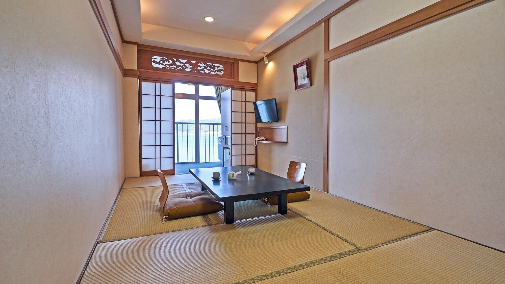 Una habitación japonesa tradicional con suelo de tatami, una mesa baja con juego de té, un televisor en la pared y un puertas correderas que dan a un balcón.
