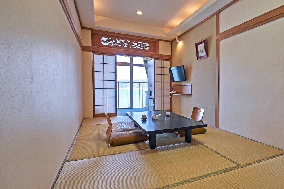Ein traditioneller japanischer Raum mit Tatami-Boden, einem niedrigen Tisch mit Teeservice, einem Fernseher an der Wand und einer Schiebetür, die auf einen Balkon führt.