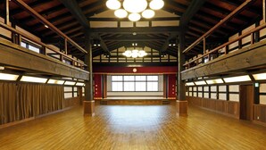 Un amplio salón con pisos de madera y techos altos, que presenta un escenario central con un fondo rojo y un ventanal grande.