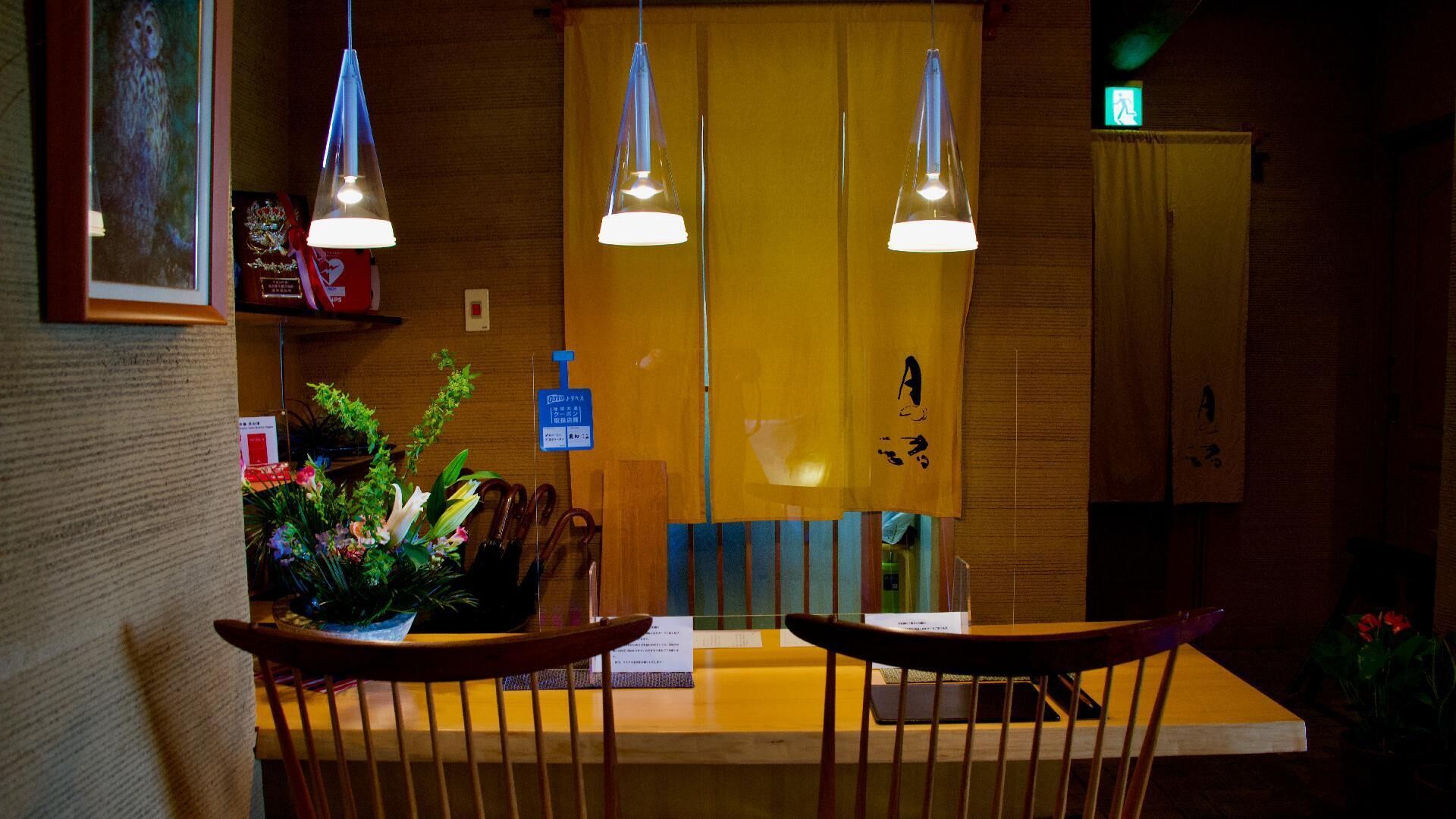 A restaurant interior with wooden chairs, hanging pendant lights, a yellow counter, and a sign with Japanese characters.