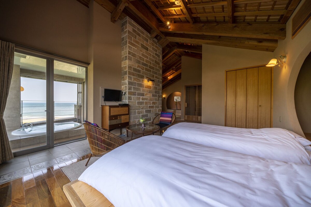 A bedroom with two beds, a stone fireplace, a television, and a view of the sea.