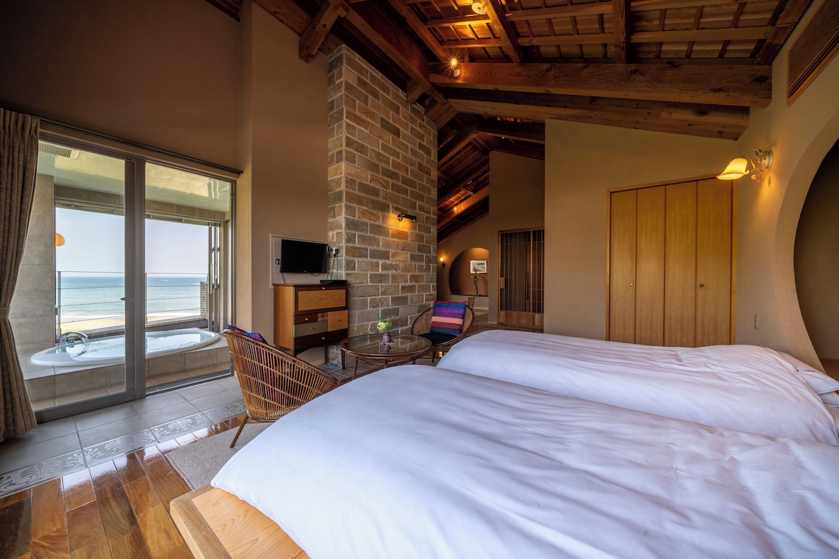 A hotel room with a large bed, a wooden ceiling, a stone fireplace, a TV, and a balcony with a view of the ocean.