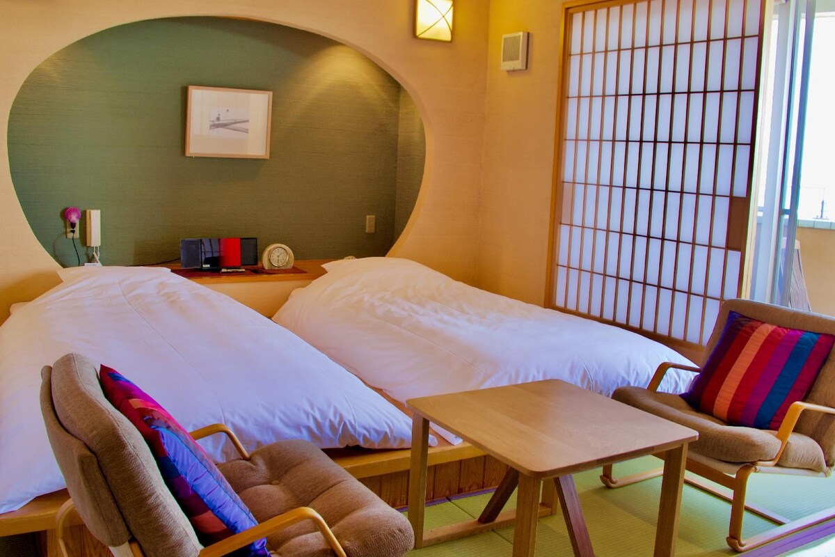 A room with two beds, a small table, and a chair with a colorful cushion.