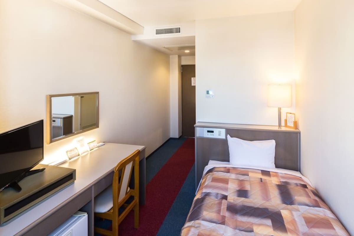 A hotel room with a bed, a desk with a computer, a chair, a television, and a lamp.