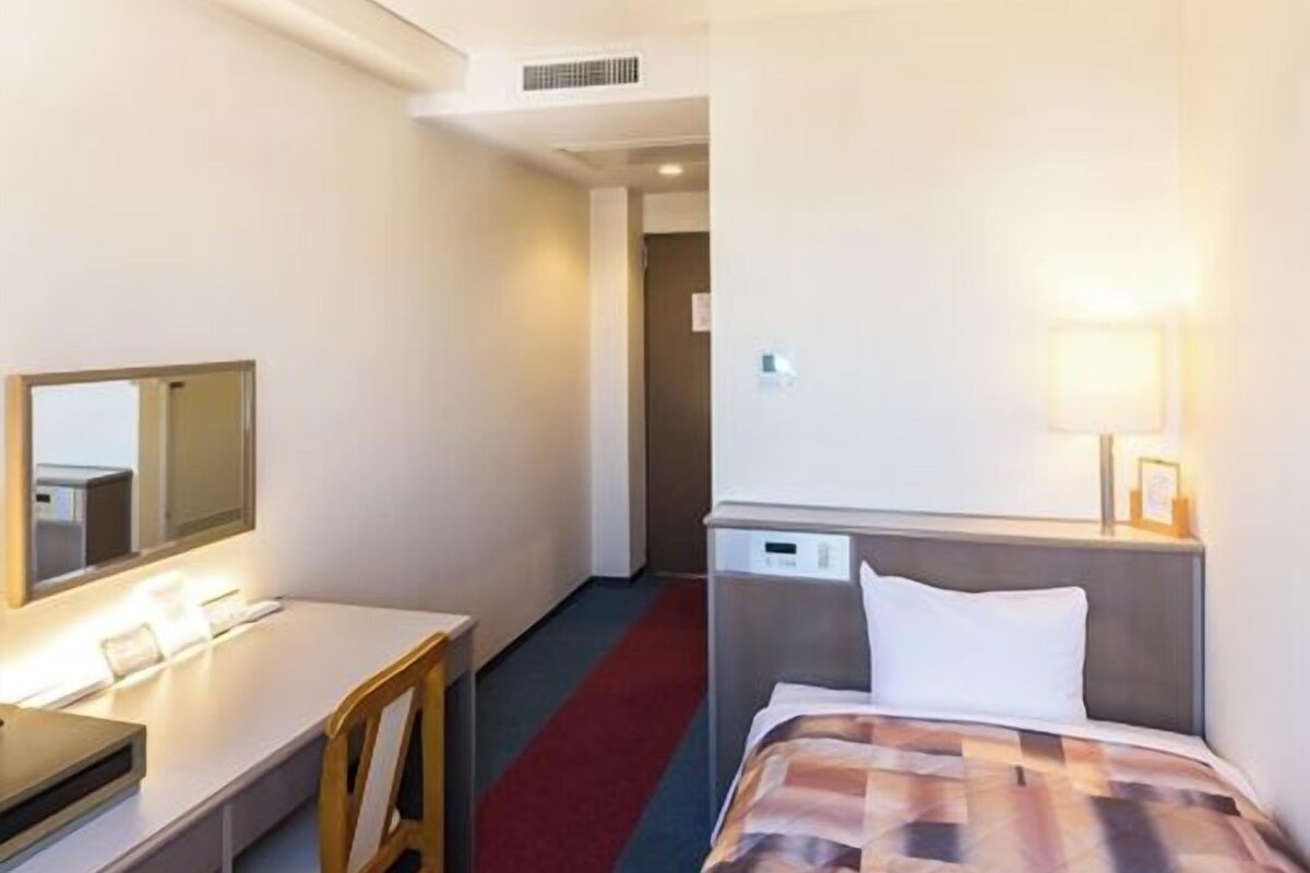 A hotel room with a bed, a desk, a chair, a mirror, and a lamp.