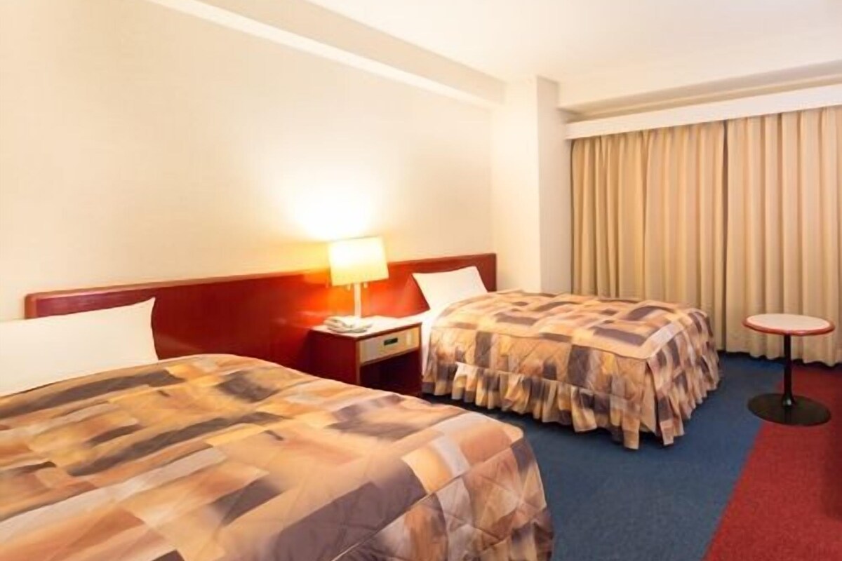 A hotel room with two beds, a nightstand with a lamp, a small table, and a window with curtains.