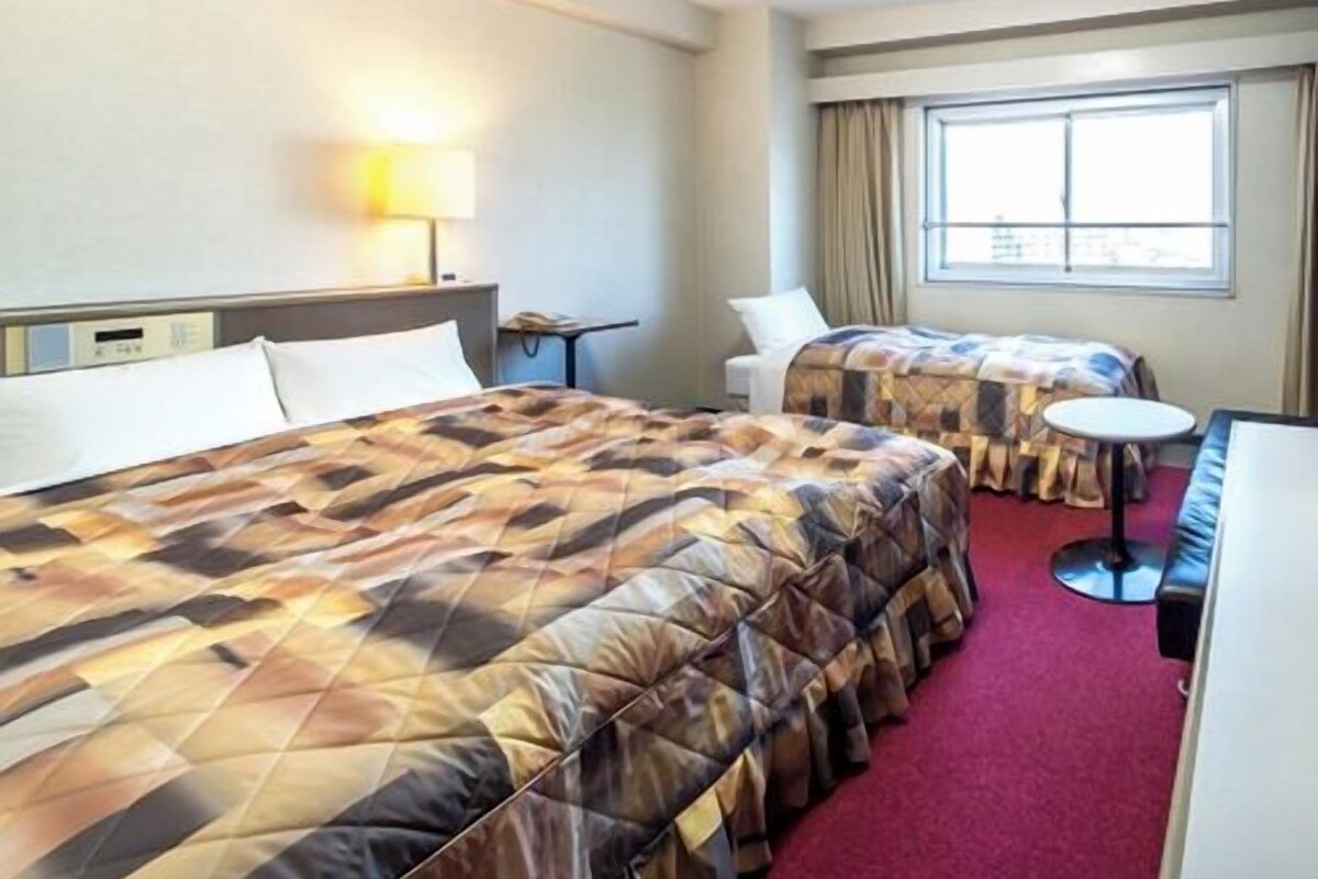 A hotel room with two beds, a desk, a chair, and a window.