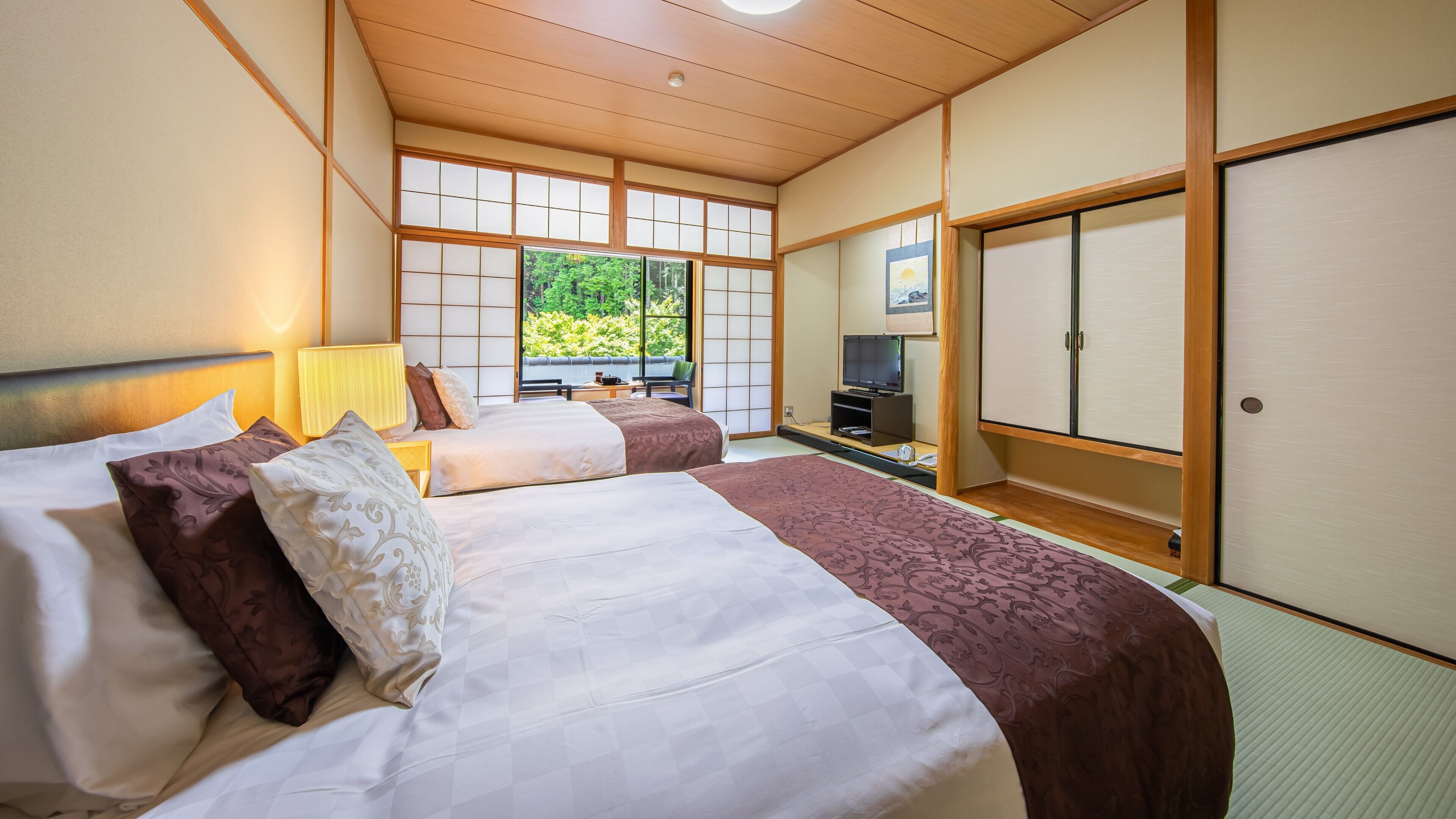 Habitación de hotel al estilo tradicional japonés, con suelo de tatami, dos camas con ropa de cama estampada, un televisor y una puerta corredera que da a un balcón.