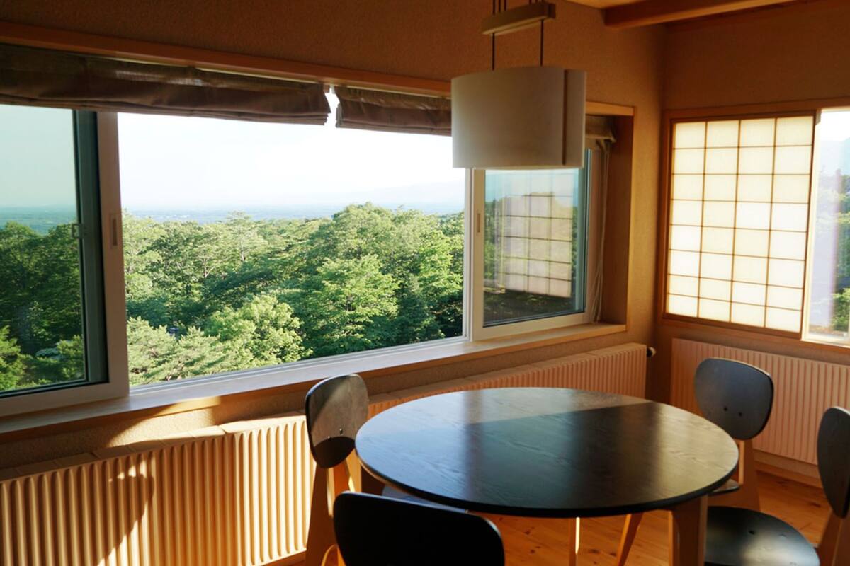 Una habitación con una mesa redonda, dos sillas, un radiador y grandes ventanales con vistas a zonas verdes.