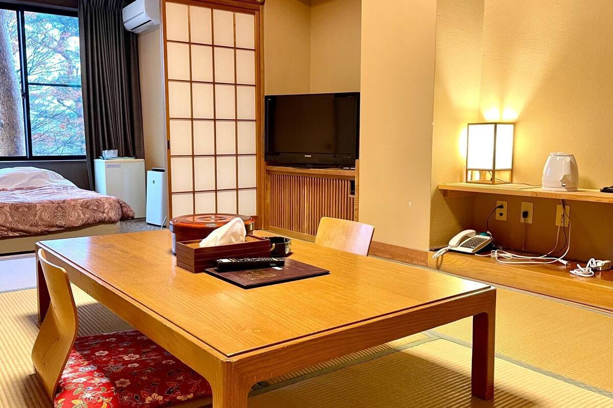 Habitación de hotel con cama, una mesa de madera, televisor, escritorio con lámpara y vistas a árboles desde la ventana.