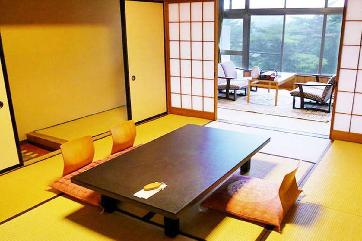 Una habitación de tatami con una mesa baja y cojines, una puerta corrediza estilo shoji y vistas a un jardín a través de amplias ventanas.