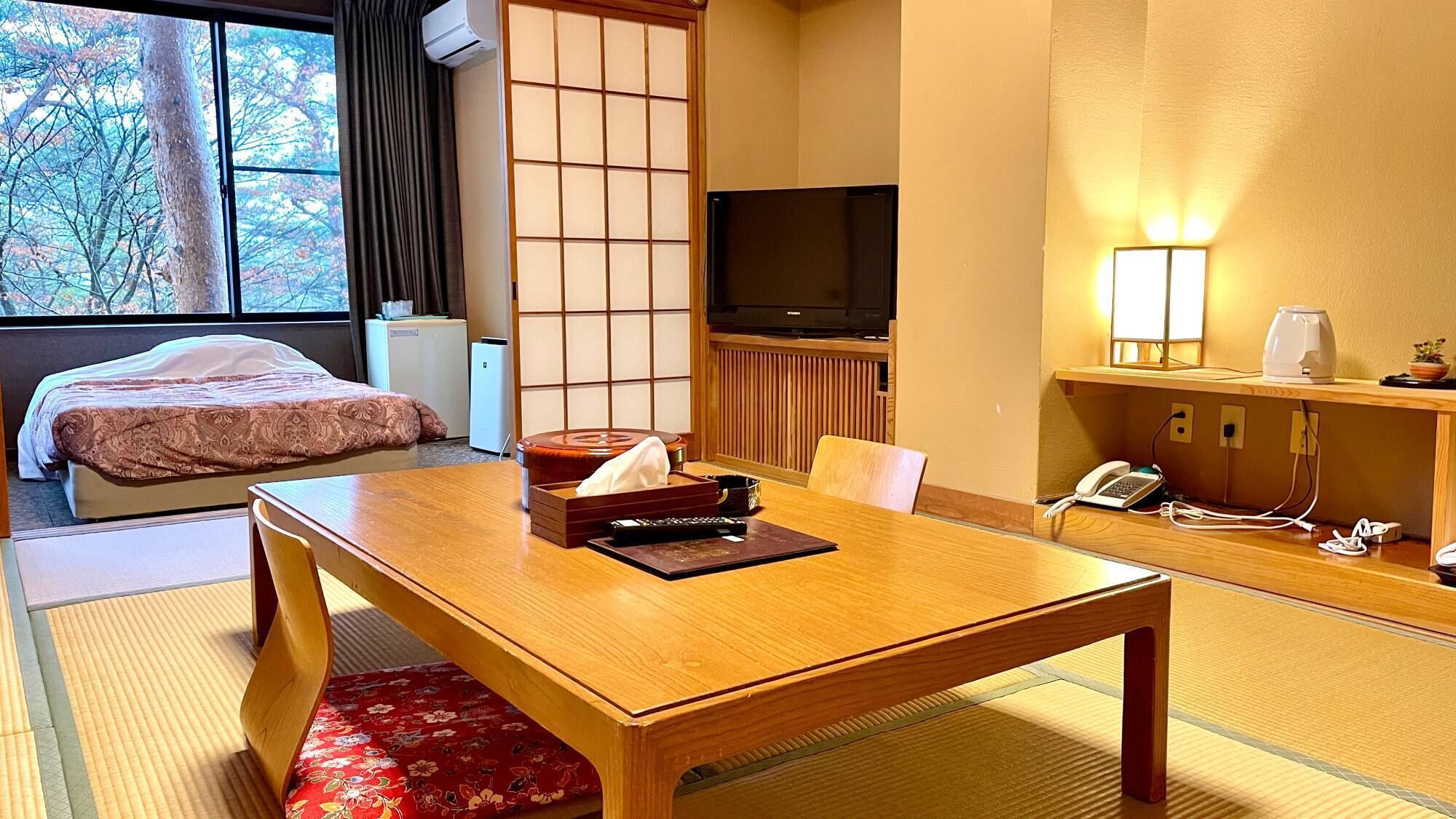 A hotel room with a bed, a wooden table, a TV, a desk with a lamp, and a view of trees through the window.