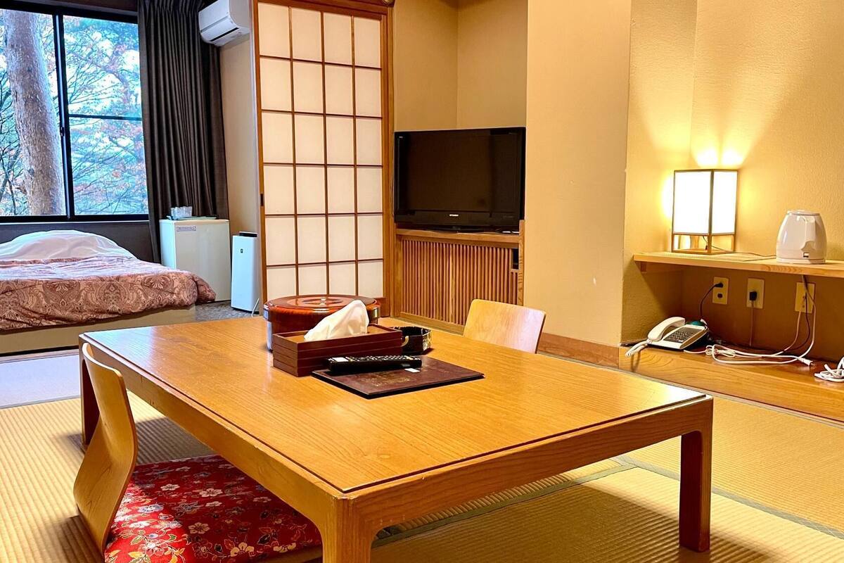 Habitación de hotel con una cama, una mesa de madera, un televisor, un escritorio con una lámpara y vistas a árboles desde la ventana.