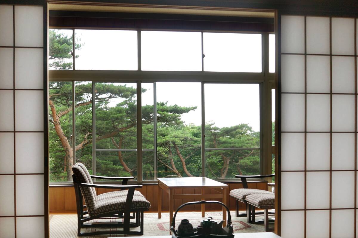 Una habitación japonesa tradicional con suelo de tatami, una mesa baja, dos sillas y vistas a árboles a través de amplios ventanales.