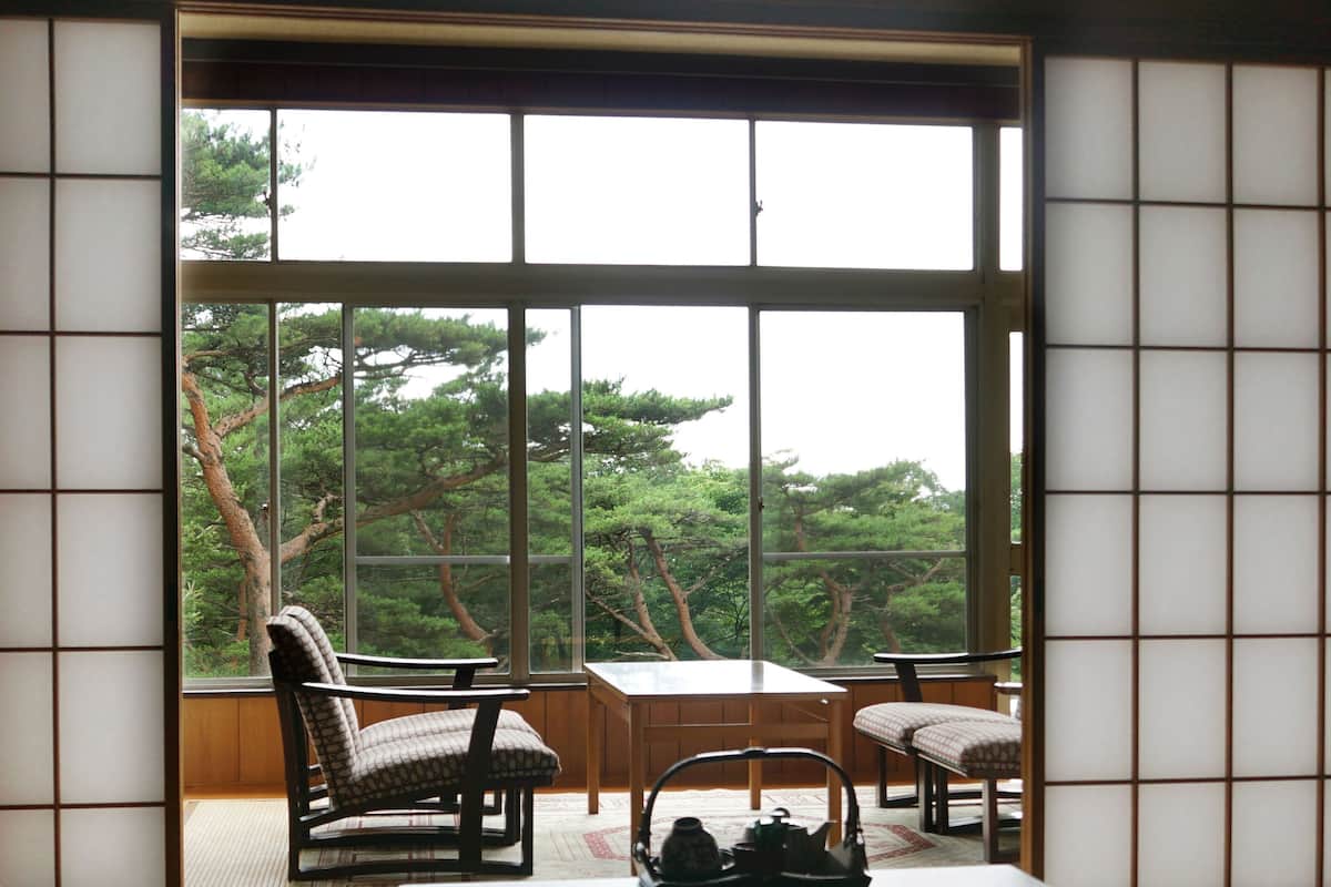A traditional Japanese room with tatami flooring, a low table, two chairs, and a view of trees through large windows.