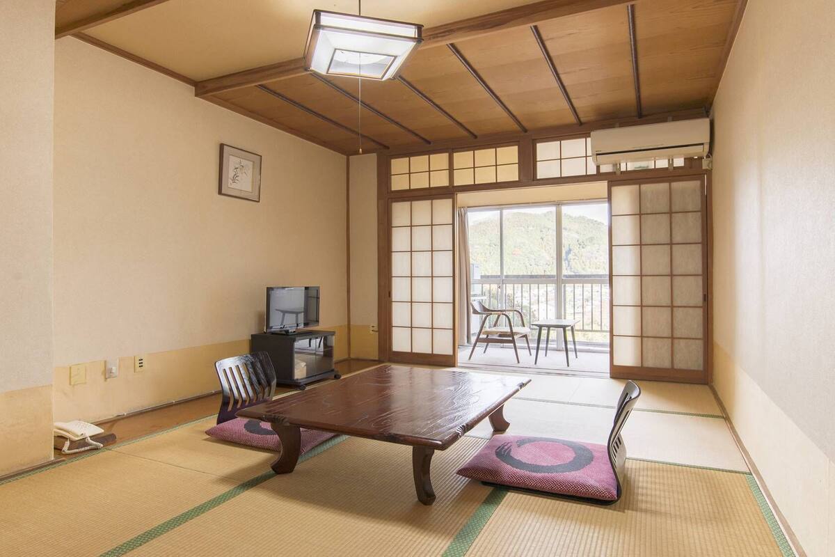 Ein traditioneller japanischer Raum mit Tatami-Böden, einem niedrigen Tisch, einem Fernseher und einer Schiebetür, die auf einen Balkon führt.
