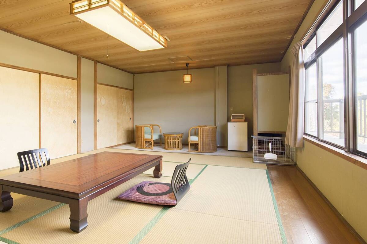 Ein traditioneller japanischer Raum mit Tatami-Bodenbelag, einem Holztisch, Stühlen, einem kleinen Schrank und einem Fenster mit Blick.