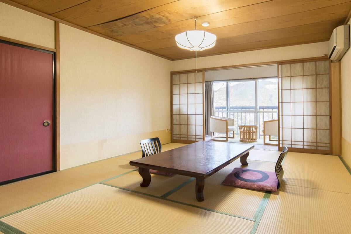 Ein Raum mit Tatami-Matten, einem Holztisch, Stühlen, einer Schiebetür und Blick durch einen Shoji-Bildschirm.