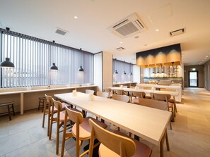A modern dining area with long tables and chairs, large windows with blinds, and pendant lighting. - Super Hotel Anan Shiyakushomae (Anan)