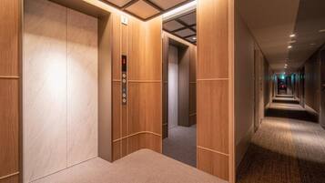 A hotel corridor with wooden panel walls, carpeted floors, and a visible elevator with a digital display.