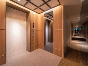 A hotel corridor with wooden panel walls, carpeted floors, and a visible elevator with a digital display.