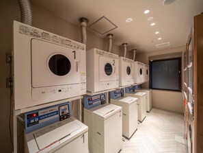 A row of white laundry machines with control panels, numbered from 1 to 5, in a hallway with a herringbone floor pattern. - Super Hotel Anan Shiyakushomae (Anan)