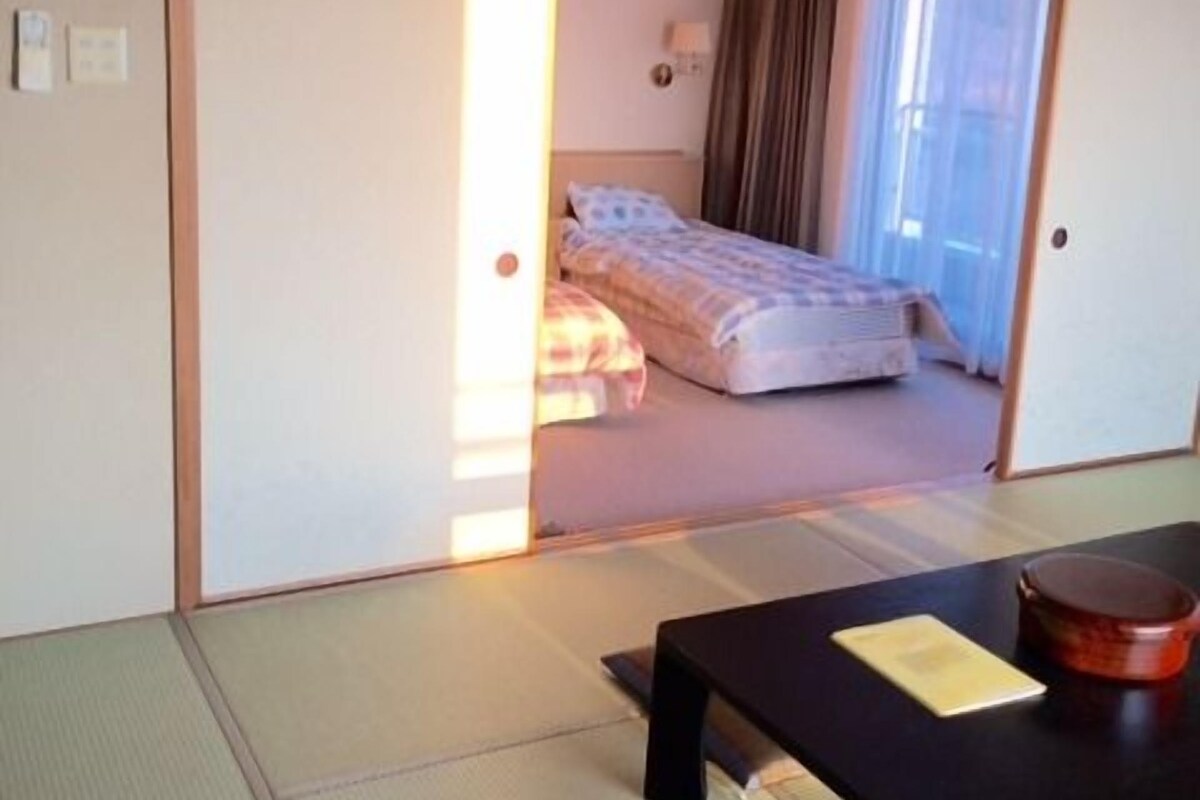 A traditional Japanese room with tatami flooring, a low table with a red container, and a bed with a blue blanket.